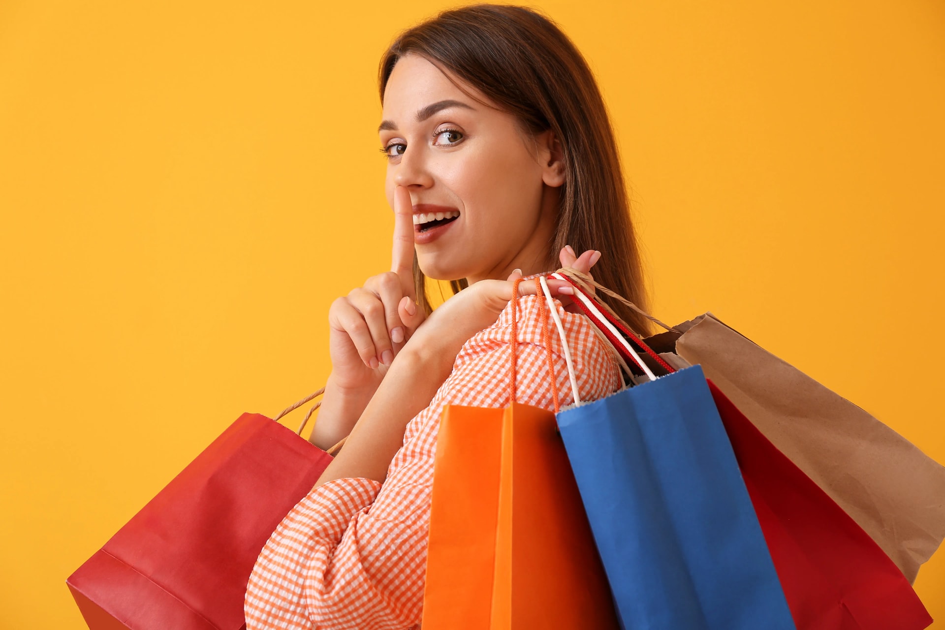 Femme-souriante-avec-des sacs-de-shopping-colorés-faisant-un-geste-de-silence-sur-fond-jaune.