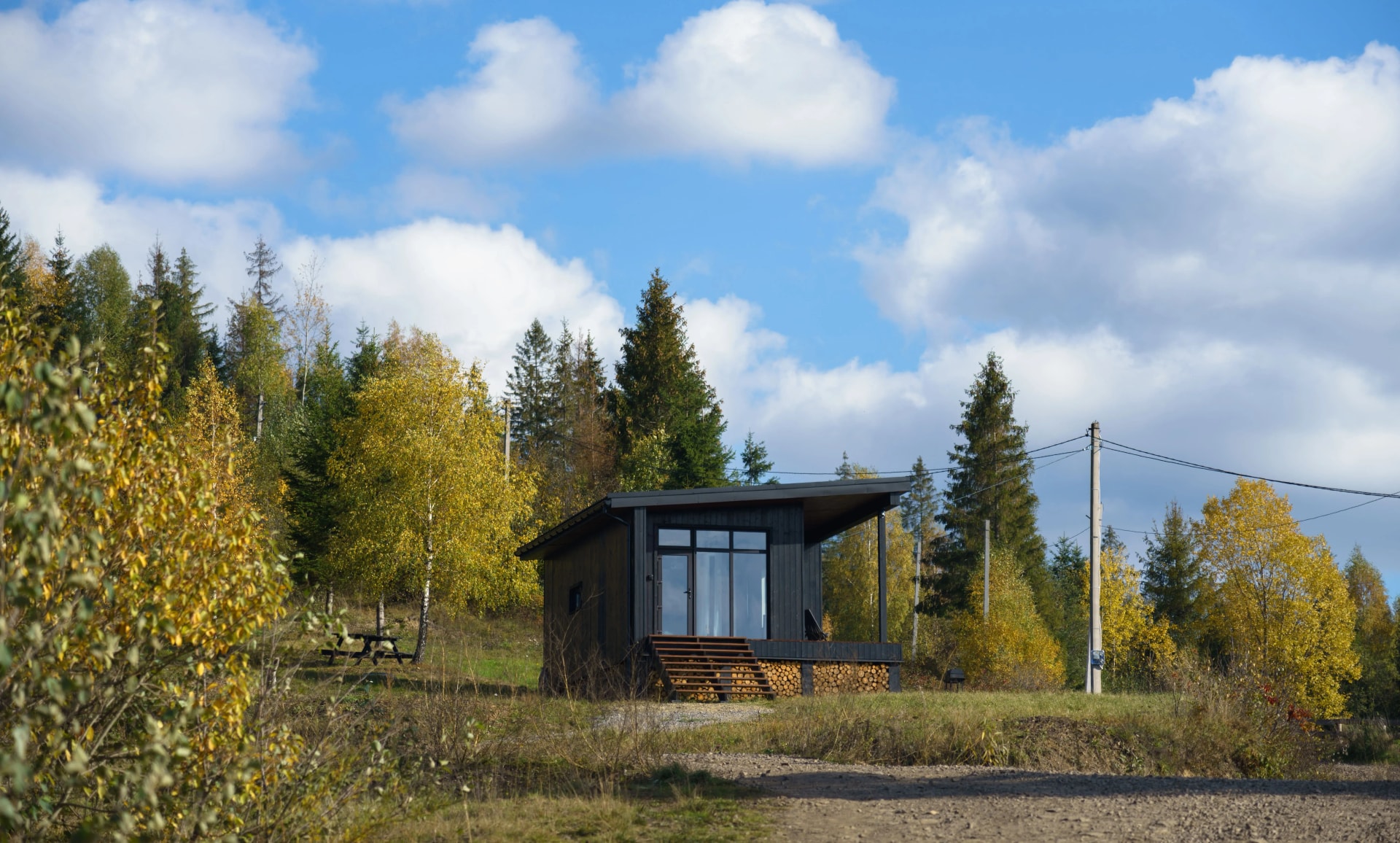 Tiny-house-en-bois-située-en-pleine-nature,-entourée-d'arbres-aux-couleurs-automnales-sous-un-ciel-bleu-avec-quelques-nuages.